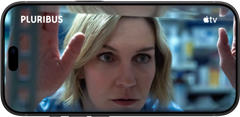 iPhone 18 Pro v orientaci na šířku, na obrazovce přiblížený výjev s ženou dívající se z okna. Název seriálu: Pluribus